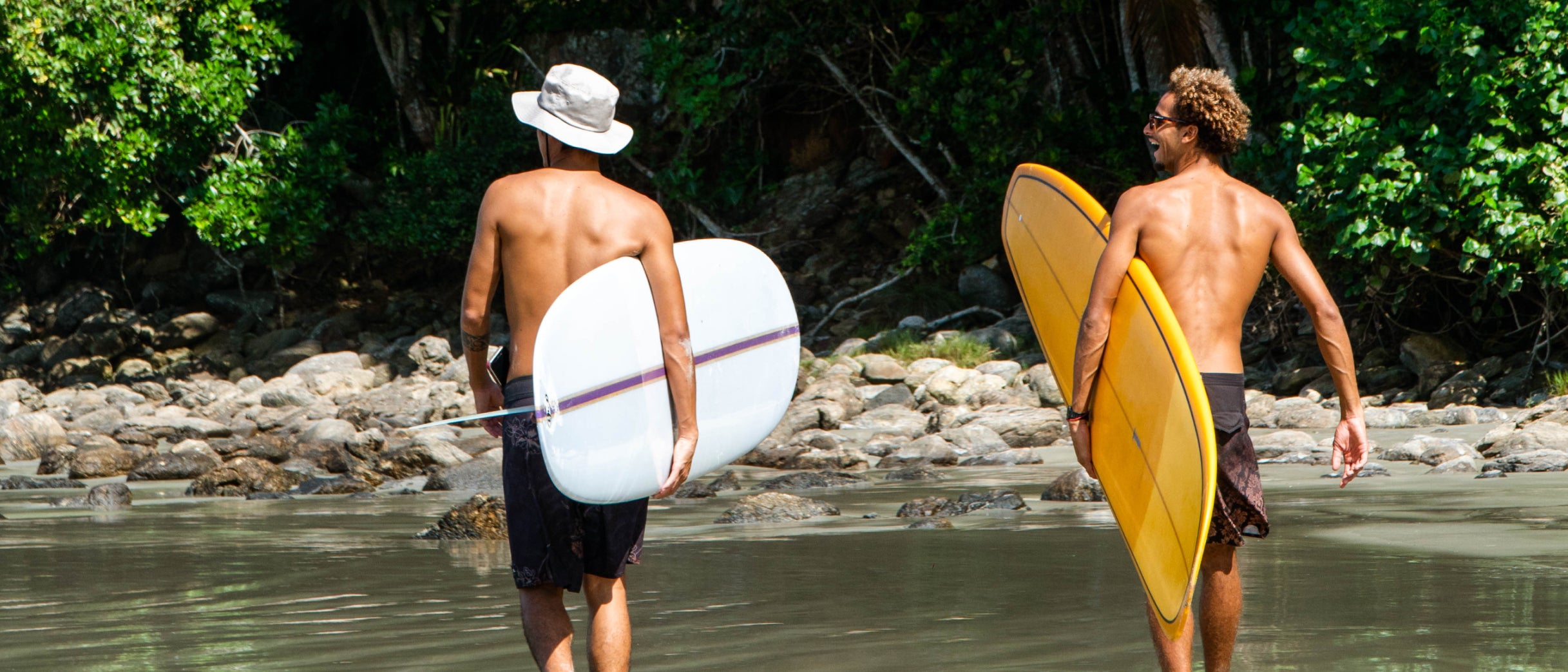 Dois surfistas caminhando em água rasa carregando pranchas de surf, cercados por pedras e vegetação tropical.